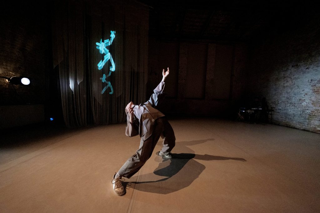 A single dancer in the foreground bending backwards on stage in a brick building.