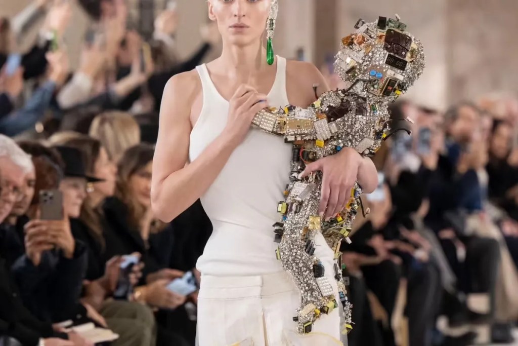 Image of model walking a fashion show carrying a baby doll encrusted with computer hardware.