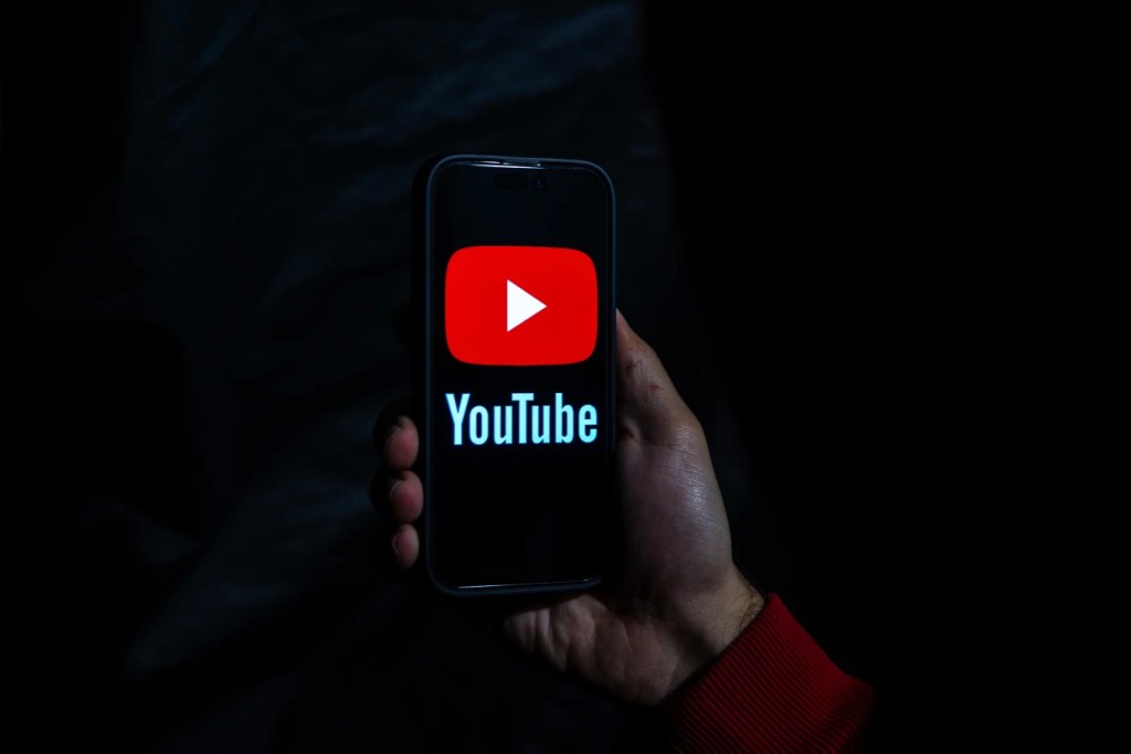 Dark background with a mobile phone in the foreground showing the YouTube logo and Play screen