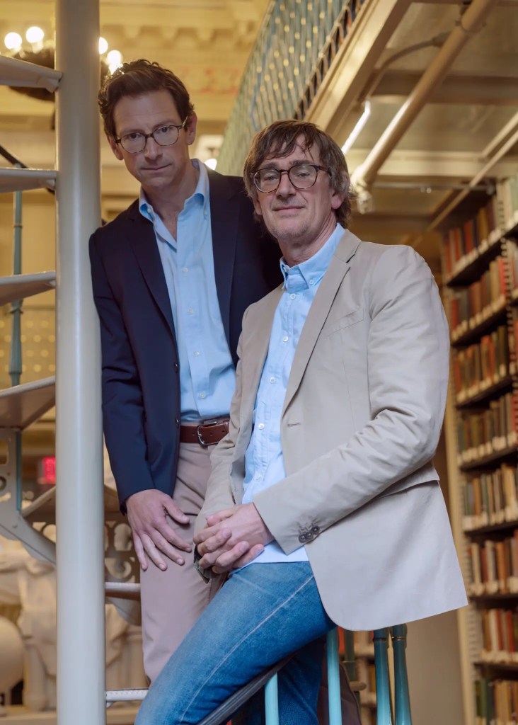 Two men in sport coats in foreground and a library and staircase in background